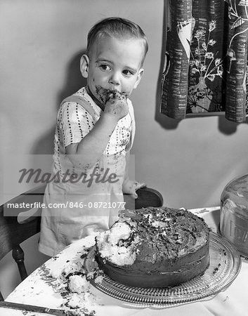 1950s LITTLE BOY TODDLER STANDING ON CHAIR BY CHOCOLATE CAKE WITH FINGERS IN MOUTH AND ICING ALL OVER HIS FACE