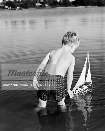 1960s YOUNG BOY WEARING BATHING SUIT STANDING IN WATER LAUNCHING TOY SAILING BOAT