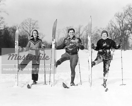 1930s TWO WOMEN AND A MAN LOOKING AT CAMERA SMILING POSING ON SKIS WITH RIGHT SKI TURNED UPRIGHT