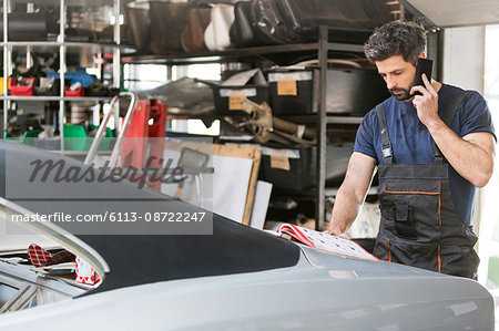 Mechanic talking on cell phone ordering car parts from catalog in auto repair shop Mechanic talking on cell phone ordering car parts from catalog in auto repair shop