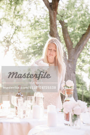 Blond woman setting a table in a garden, candles and vases with pink roses.
