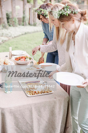 Women with flower wreath in their hair at a garden party, choosing food from a buffet.