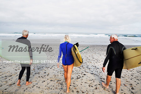 Senior woman and two senior men on a beach, wearing wetsuits and carrying surfboards.