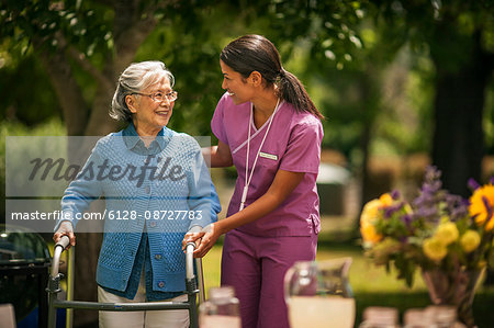 Friendly young nurse helps an elderly lady exercise with her walker in a sunny garden.