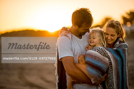 Happy couple cuddle their young daughter on a beach at sunset.