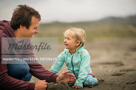 Young girl builds sandcastles with her father on a gray day at the beach.