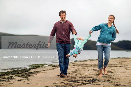 Affectionate young couple swing their little girl between them on a walk at the beach.