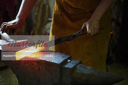 A blacksmith holds a length of red hot metal on an anvil with a pair of tongs.