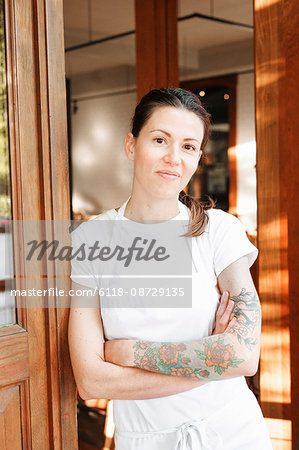 Portrait of a woman wearing a white apron standing with her arms crossed in a doorway, tattoos on her left arm.