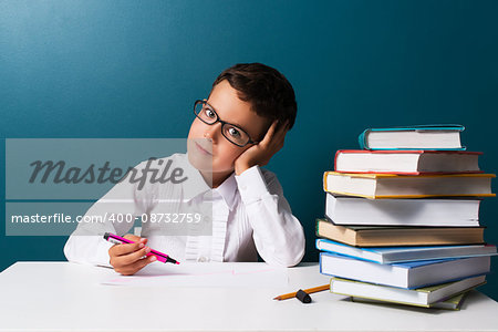 Pensive cute boy with glasses sitting at a table, blue background
