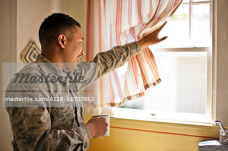 Smiling young adult soldier holding a mug while looking out the window of his home.