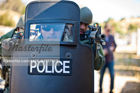 Military policeman aiming guns from behind a police riot shield.