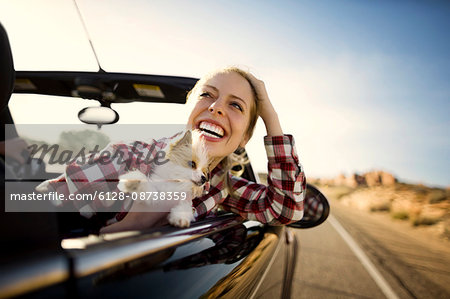 Laughing young woman sitting in the passenger seat of a convertible car with her small dog on a road trip.