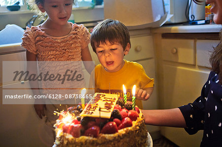 Young boy blowing out his birthday candles as his smiling mother holds the birthday cake in their kitchen.