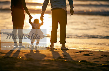 Young parents support their baby daughter between them as she takes her first steps on the beach at sunset.