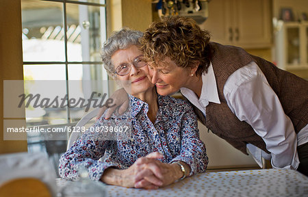 Smiling mature woman greets her cheerful elderly mother as she sits at the kitchen table.