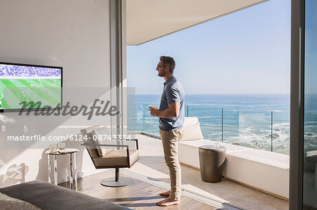 Man watching soccer on TV at sunny luxury patio doorway with ocean view