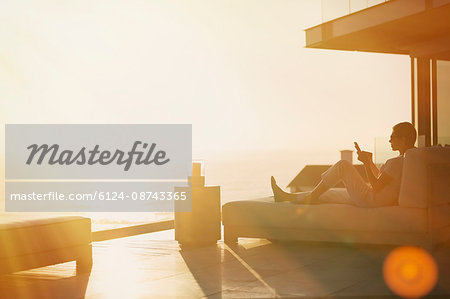 Silhouette woman using cell phone on chaise lounge on luxury balcony with sunset ocean view