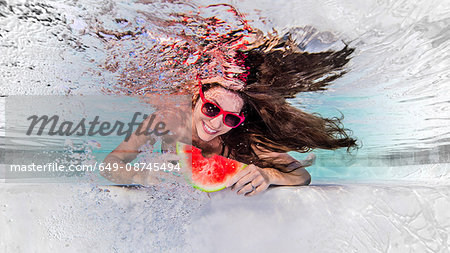 Underwater view of woman wearing sunglasses, eating slice of water melon, smiling