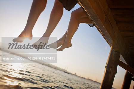 Two pairs of legs dangling off a wooden pier.