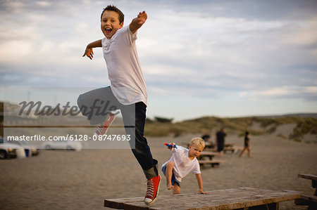 Two boys jumping from picnic tables at the beach.