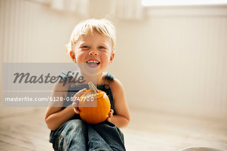 Young boy holds a small Jack O'Lantern on his knees as he sits on a floor and poses for a portrait.