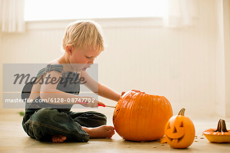 Young boy sitting cross-legged on the floor leans forward to carve the face of a Jack O'Lantern with a knife on a big pumpkin with a finished small Jack O'Lantern next to him.