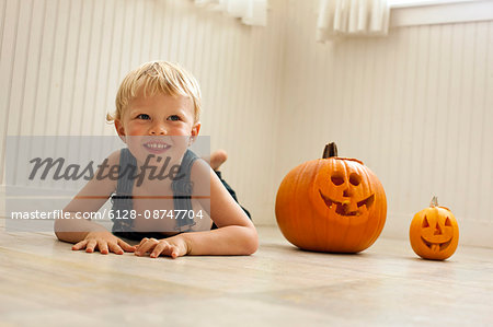 Smiling,  young boy lies front down on a floor next to a big Jack O'Lantern and a small Jack O'Lantern as he poses for a portrait.