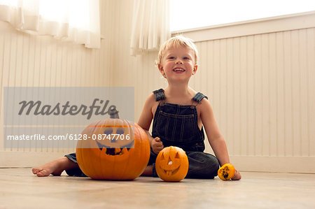 Smiling,  young boy sits a floor behind a big Jack O'Lantern and a small Jack O'Lantern as he poses for a portrait.
