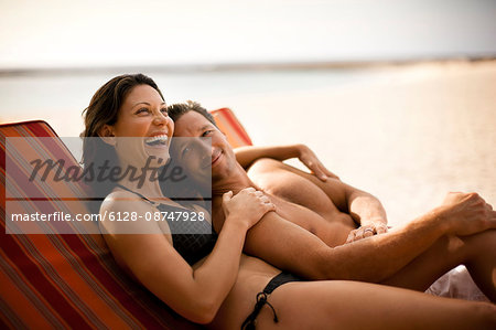 Happy mid-adult couple laughing while lying side by side on a deck chair at a beach resort.