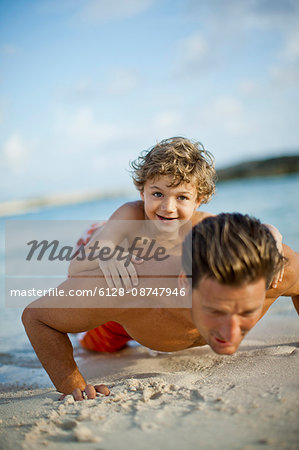 Portrait of a young boy lying on his fathers back as he does press ups on a sandy beach.