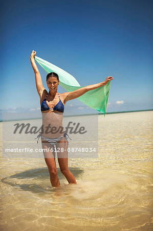 Portrait of a happy young woman lifting her towel above her head as she enjoys wading in the tropical water.