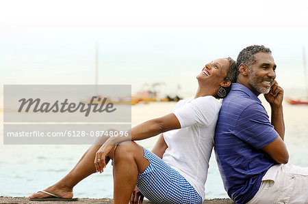 Happy mature couple sitting back to back by the beach.