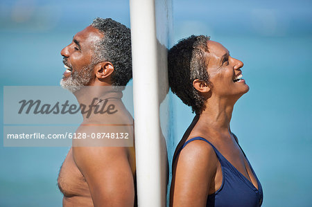 Laughing mature couple standing back to back on a beach.