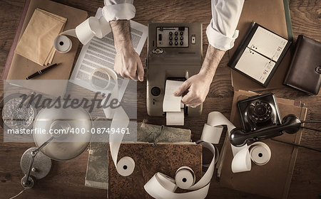 Messy vintage accountant's desktop with adding machine and paper rolls, he is working with the calculator