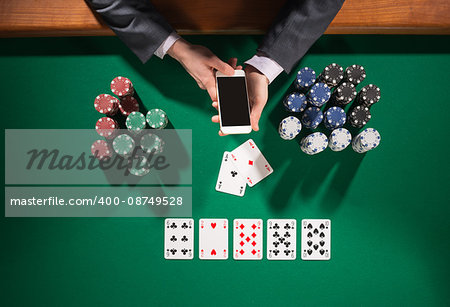 Elegant male poker player using a smartphone, cards and chips stacks all around, green table top view