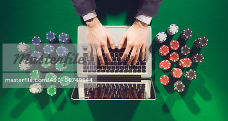 Online player's hands with laptop and stack of chips all around on green table top view, he is typing on the keyboard