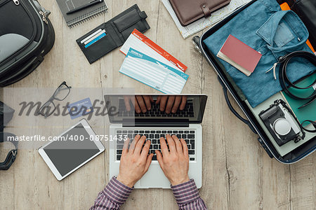 Traveler getting ready for a trip, he is packing his bag and planning a journey online using a laptop, flat lay