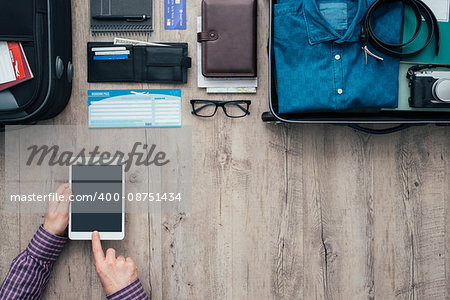 Man packing for a trip, he is using a digital touch screen tablet and booking flights online using a mobile app, flat lay banner