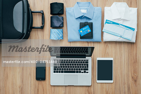 Planning a business trip: bag, formal clothing, credit cards and plane tickets on a desk with laptop, smartphone and tablet, traveling and technology concept