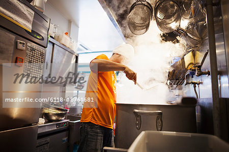 The ramen noodle shop kitchen. A vast pot on the stove, and steam rising.  A man cooking noodles..