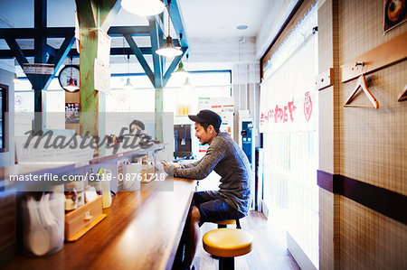 A ramen noodle shop in a city. A man seated at a counter looking at his smart phone.