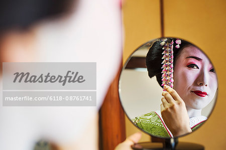 A woman made up in the traditional geisha style, with an elaborate hairstyle and floral hair clips, with white face makeup with bright red lips and dark eyes. Looking in a hand mirror.