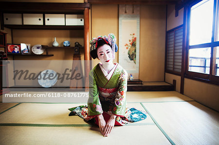 A woman dressed in the traditional geisha style, wearing a kimono and obi, with an elaborate hairstyle and floral hair clips, with white face makeup with bright red lips and dark eyes, kneeling in a traditional pose.