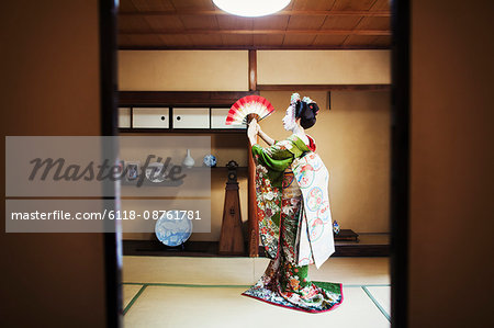 A woman dressed in the traditional geisha style, wearing a kimono and obi, with an elaborate hairstyle and floral hair clips, with white face makeup holding an open fan up, her kimono arranged on the floor.