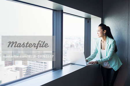 A business woman by a window with a view over the city, looking out. Laptop.
