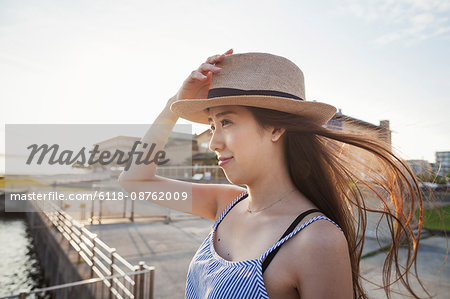 Young woman standing on a pier by water holding her straw hat on her head.