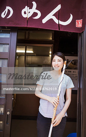 Woman standing by the door of a small noodle shop with a brush.