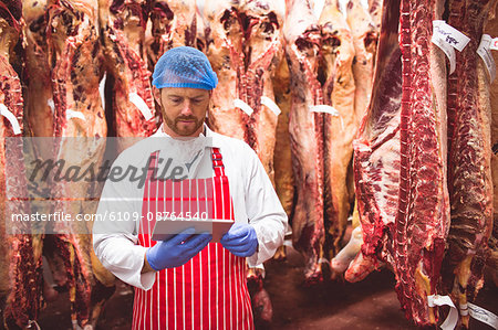 Butcher using digital tablet in meat storage room at butchers shop