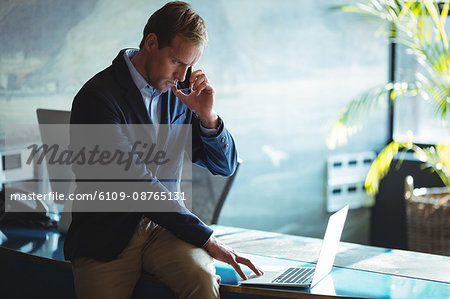 Businessman talking on mobile phone while using laptop in office